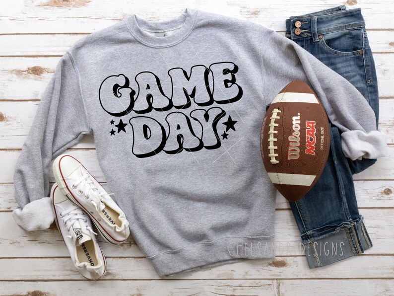 Op de afbeelding: Een grijs sweatshirt met de tekst "GAME DAY" in zwart, gestileerd lettertype. Een bruine voetbal met witte strepen en de tekst "Wilson NCAA" bevindt zich aan de rechterkant van de afbeelding.