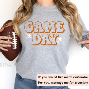 Op de afbeelding: Een grijs sweatshirt met de tekst "GAME DAY" in oranje retro-lettertype. De tekst wordt omringd door witte sterren. Een persoon houdt een bruine voetbal vast.