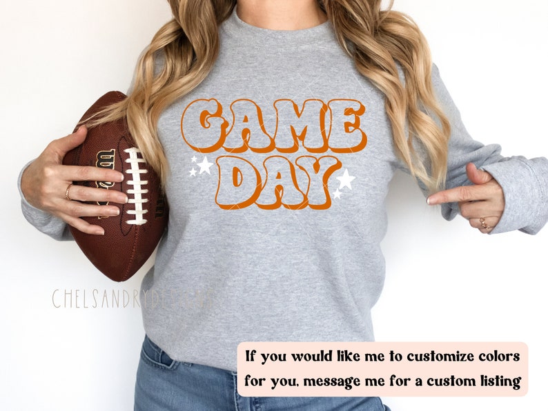 Op de afbeelding: Een grijs sweatshirt met de tekst "GAME DAY" in oranje retro-lettertype. De tekst wordt omringd door witte sterren. Een persoon houdt een bruine voetbal vast.