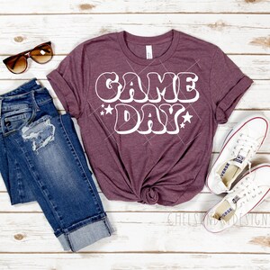 Op de afbeelding: Een maroon t-shirt met de tekst "GAME DAY" in wit, omrand met zwart, met witte sterren aan weerszijden van de tekst. Het shirt ligt op een wit houten oppervlak met een blauwe jeans, witte sneakers en een zonnebril.