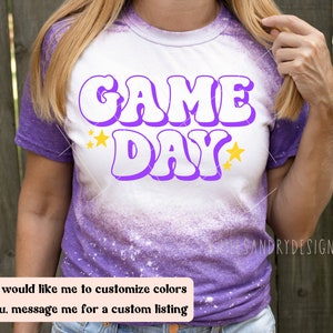 Op de afbeelding: Een wit T-shirt met een paarse "GAME DAY" afbeelding met gele sterren. Het shirt is in tie-dye stijl met paars en wit.