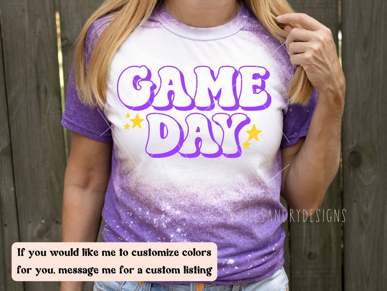 Op de afbeelding: Een wit T-shirt met een paarse "GAME DAY" afbeelding met gele sterren. Het shirt is in tie-dye stijl met paars en wit.