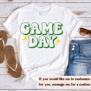 Op de afbeelding: Wit T-shirt met groene en gele tekst "GAME DAY" met gele sterren aan weerszijden van de tekst.