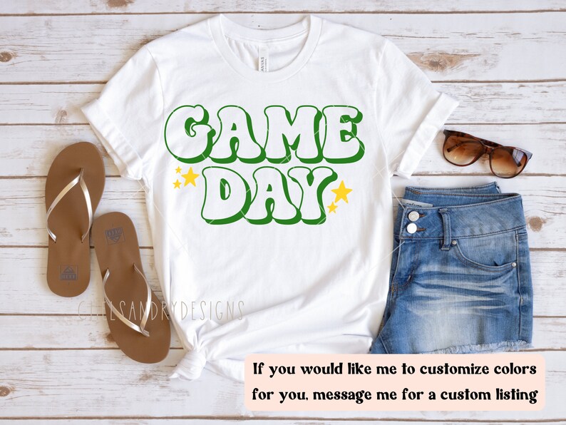 Op de afbeelding: Wit T-shirt met groene en gele tekst "GAME DAY" met gele sterren aan weerszijden van de tekst.