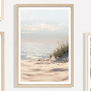 May include: Framed beach scene print featuring a sandy shore, ocean, and sky. The artwork showcases a serene coastal landscape with soft colors, including beige, blue, and white. The print is in a light-colored wooden frame, suitable for home decor.
