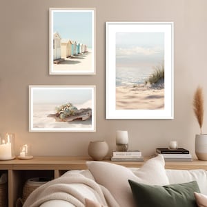 May include: Three framed art prints with beach themes. One print shows colorful beach huts, another depicts a sandy beach with a succulent, and the third displays a serene ocean view with sand and grass. The frames are light-colored.