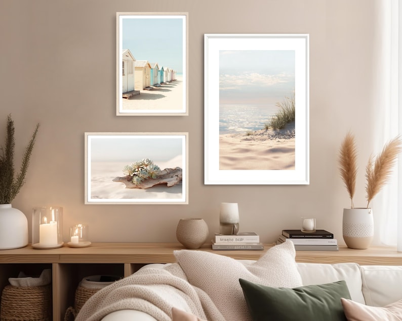 May include: Three framed art prints with beach themes. One print shows colorful beach huts, another depicts a sandy beach with a succulent, and the third displays a serene ocean view with sand and grass. The frames are light-colored.