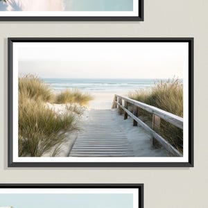 May include: A framed print featuring a beach scene. A wooden boardwalk leads to the ocean, flanked by tall, golden grasses. The sky is a soft, muted blue, and the sand is white.