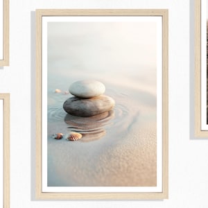 May include: A framed print featuring a serene beach scene. Two smooth stones are stacked on top of each other in the water, with seashells scattered around. The image has a soft, natural color palette with warm tones.