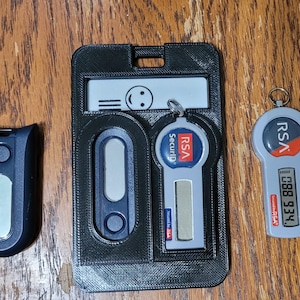 May include: An assortment of security tokens and a holder, displayed on a wooden surface. The items include a black token, a black holder with a token and a keychain token with the text "RSA SecurID".