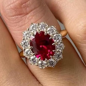May include: A gold ring featuring a large, oval-cut, deep red gemstone surrounded by a halo of smaller, clear gemstones. The ring's band is a simple, polished gold.