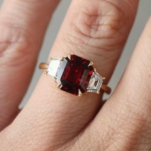 Puede incluir: Un anillo de oro con una gran gema rectangular de color rojo intenso, flanqueada por dos piedras blancas más pequeñas en forma de trapecio. El anillo está en un dedo, mostrando el diseño y el contraste de color de la joya.
