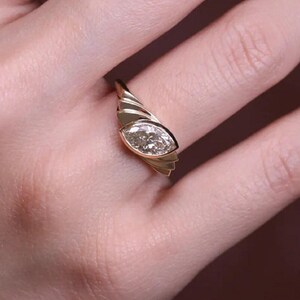 May include: A gold ring with a marquise-cut diamond. The ring features a unique design with a textured band that wraps around the finger. The diamond is set in a bezel setting, and the ring is shown on a hand.