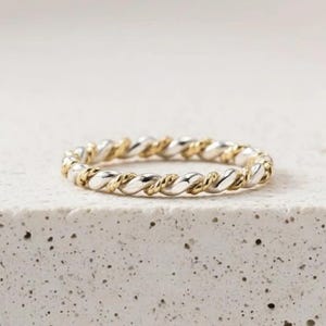 May include: A two-tone ring with a twisted rope design. The ring features alternating sections of gold and silver metal. The ring is displayed on a white, textured surface. The ring is a delicate piece of jewelry.