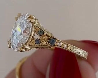 Pode incluir: Um anel de ouro com uma grande pedra central de corte oval e uma banda com detalhes intrincados em filigrana. A cravação inclui pequenas pedras de destaque azul-escuras embutidas no metal ao longo dos lados da gema central.