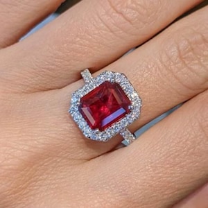 May include: A silver ring with a large, rectangular, deep red gemstone, encircled by smaller, clear gemstones. The ring is displayed on a hand, highlighting the jewellery's design and colour. The ring's design is elegant.