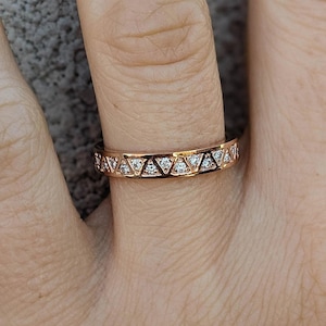 May include: Rose gold ring with a repeating geometric design. The ring features small, clear gemstones set within the triangular shapes. The ring is worn on a finger, showcasing its delicate details and sparkle.