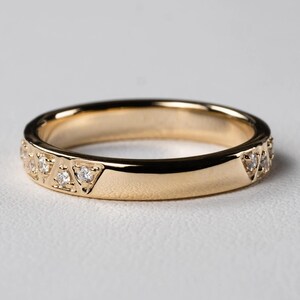 May include: A gold band ring with a polished finish. The ring features a series of small, clear gemstones set in triangular patterns along the band. The ring is shown on a white surface.