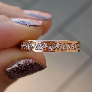 May include: A rose gold ring with a repeating pattern of triangles, each containing a small, clear gemstone. The ring is held up, showcasing the intricate design and the sparkle of the gemstones. The ring's band is a warm, metallic color.