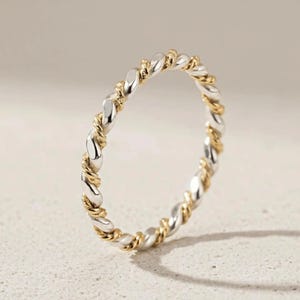 May include: A silver and gold-tone twisted ring. The ring features a repeating pattern of silver and gold-colored metal, creating a braided or rope-like design. The ring is set against a neutral background.