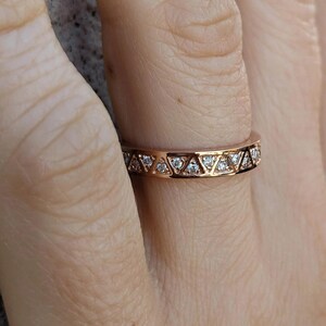 May include: Rose gold ring with a geometric design. The ring features a series of triangles, each set with a small, clear gemstone. The band is a warm, metallic color, and the gemstones sparkle.
