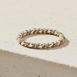 May include: A silver and gold-toned ring with a twisted rope design. The ring is circular and sits on a light-colored surface. The ring's design features alternating silver and gold segments, creating a textured appearance.