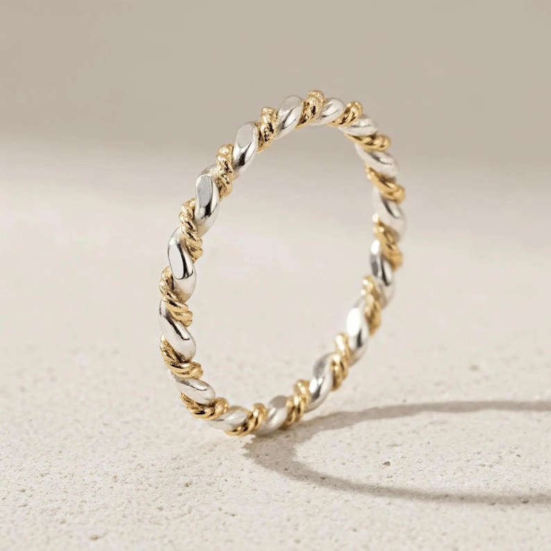 May include: A silver and gold-tone twisted ring. The ring features a repeating pattern of silver and gold-colored metal, creating a braided or rope-like design. The ring is set against a neutral background.