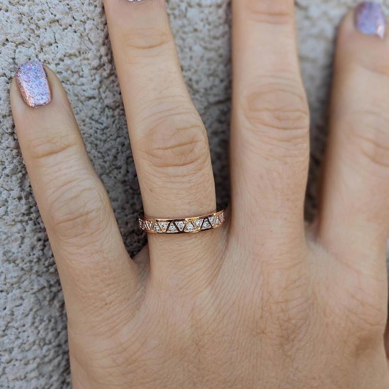 May include: Rose gold ring with a repeating geometric design. The ring features small, clear gemstones set within the triangular shapes. The ring is worn on a finger, showcasing its delicate details and sparkle.