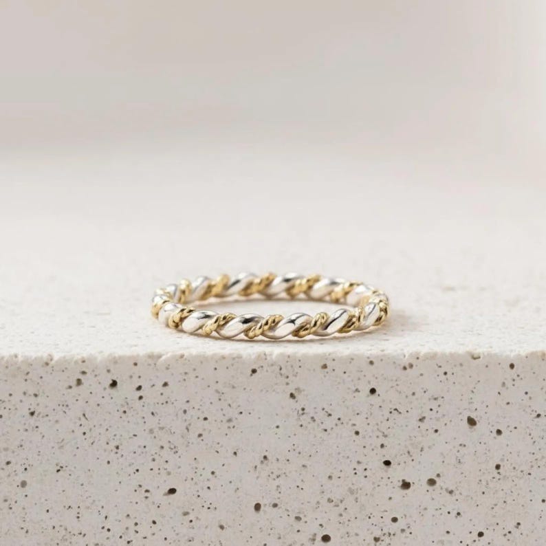 May include: A two-tone ring with a twisted rope design. The ring features alternating sections of gold and silver metal. The ring is displayed on a white, textured surface. The ring is a delicate piece of jewelry.