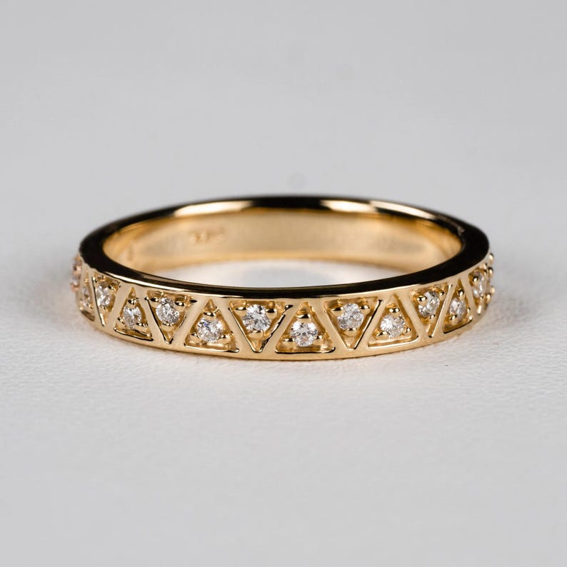 May include: A gold band ring with a repeating pattern of triangles, each containing a small, clear gemstone. The ring has a polished finish and is set against a white background. The ring is a delicate piece of jewelry.
