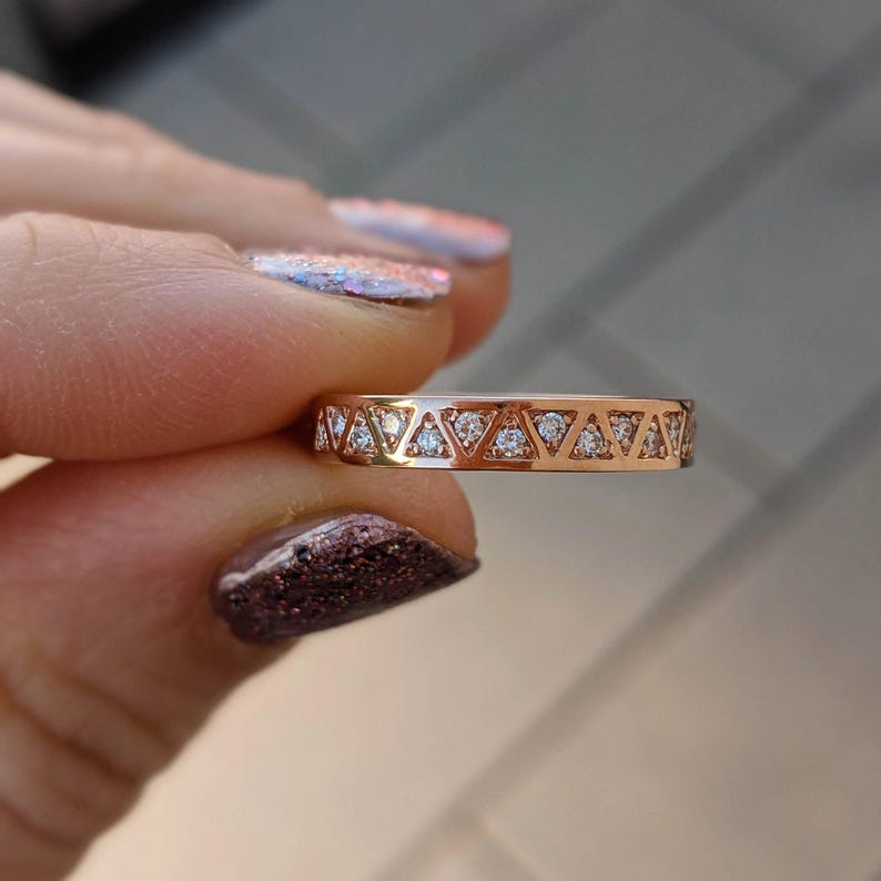 May include: A rose gold ring with a repeating pattern of triangles, each containing a small, clear gemstone. The ring is held up, showcasing the intricate design and the sparkle of the gemstones. The ring's band is a warm, metallic color.