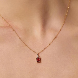 Puede incluir: Un collar dorado con un colgante rectangular de piedra preciosa rojo oscuro. El colgante está acentuado con una pequeña piedra transparente. La delicada cadena tiene pequeñas cuentas espaciadas uniformemente.