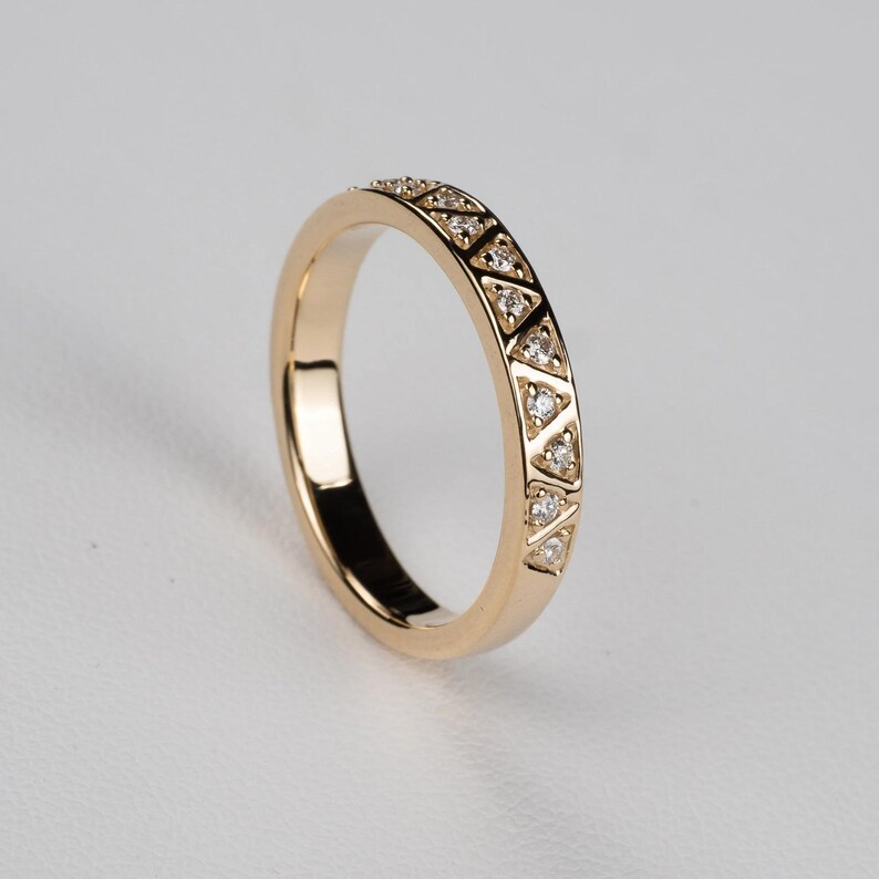 May include: A gold ring with a repeating pattern of small triangles, each containing a small, clear stone. The ring has a smooth, polished band and a bright, reflective surface. The ring is a piece of jewelry.