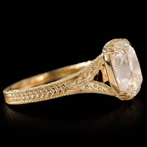 May include: A gold ring with an engraved band and a large, clear, cushion-cut gemstone. The ring's setting has intricate filigree details, and the band features a textured, leaf-like pattern. The ring is set against a black background.