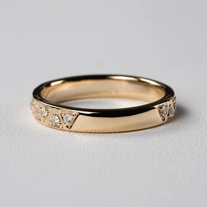 May include: A gold band ring with a polished finish. The ring features a series of small, clear gemstones set in triangular patterns along the band. The ring is shown on a white surface.