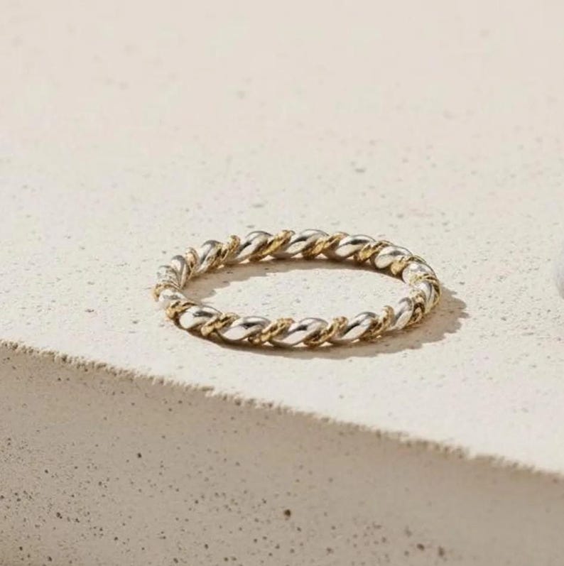 May include: A silver and gold-toned ring with a twisted rope design. The ring is circular and sits on a light-colored surface. The ring's design features alternating silver and gold segments, creating a textured appearance.