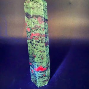Op de afbeelding: Een hoge, groene en rode unakiet kristallen obelisk. De gepolijste steen heeft een zeshoekige vorm en is bespikkeld met zwarte en rode vlekken. De bovenkant is puntig en de basis is plat. De achtergrond is donkerblauw.