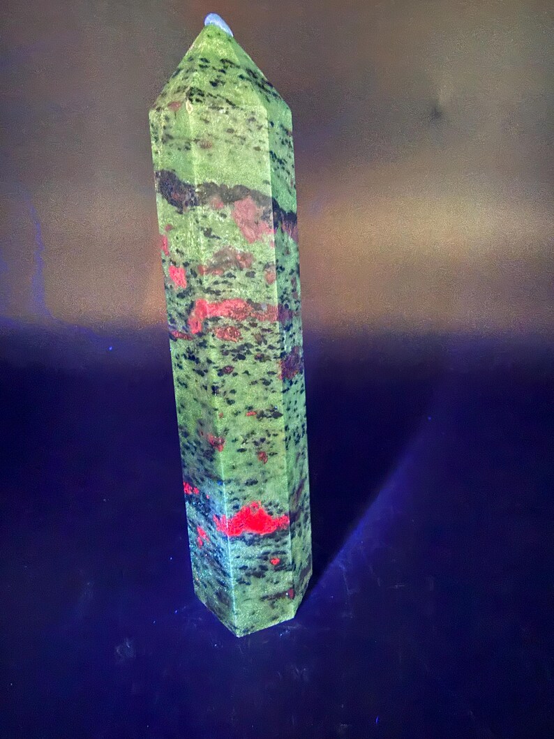 Op de afbeelding: Een hoge, groene en rode unakiet kristallen obelisk. De gepolijste steen heeft een zeshoekige vorm en is bespikkeld met zwarte en rode vlekken. De bovenkant is puntig en de basis is plat. De achtergrond is donkerblauw.