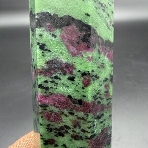 Op de afbeelding: Een rechthoekige, gepolijste edelsteen met een levendige groene basiskleur, bespikkeld met zwarte en dieprode insluitsels. Het oppervlak van de steen is glad en toont de natuurlijke patronen en texturen van het mineraal. De edelsteen is een decoratief item.