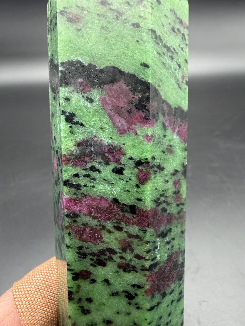 Op de afbeelding: Een rechthoekige, gepolijste edelsteen met een levendige groene basiskleur, bespikkeld met zwarte en dieprode insluitsels. Het oppervlak van de steen is glad en toont de natuurlijke patronen en texturen van het mineraal. De edelsteen is een decoratief item.