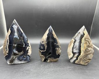 May include: Three polished agate stone sculptures with teardrop shapes. The stones feature a mix of black, white, and tan colors with intricate patterns. Each sculpture has a flat base and a pointed top, showcasing natural stone formations.