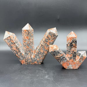 May include: Two granite crystal sculptures with a red, black, and grey speckled pattern. The sculptures are in a starburst shape, with multiple pointed tips. The crystals are on a dark grey surface.