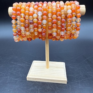 May include: A collection of multiple stretch bracelets made from round, polished carnelian beads in various shades of orange, red, and translucent white. The bracelets are displayed on a light-colored wooden T-bar stand against a dark background.