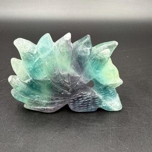 May include: A carved fluorite fox sculpture in shades of green, blue, and purple. The fox is depicted with multiple tails and a detailed, textured surface. The sculpture is displayed on a dark surface.