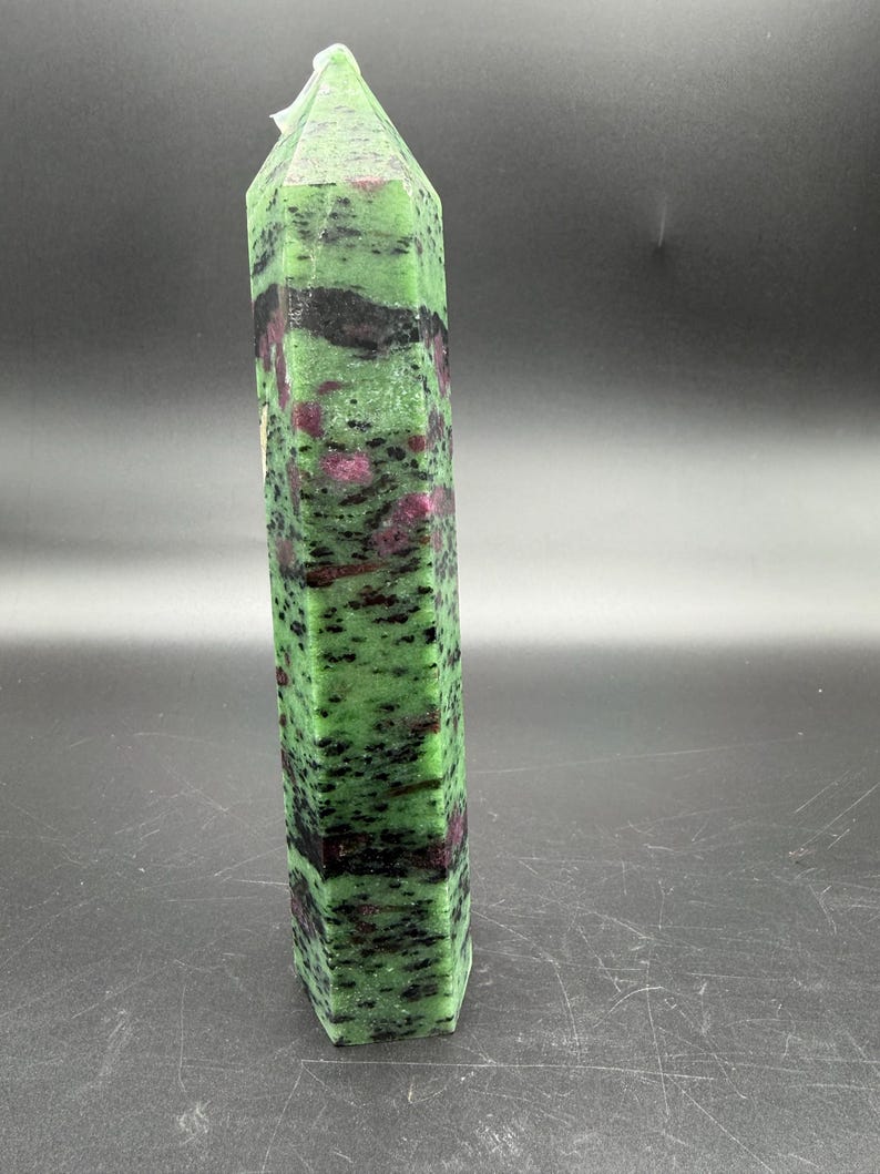 Op de afbeelding: Een hoge, gefacetteerde zoisietkristal met een groene basiskleur, bespikkeld met zwarte en rode insluitsels. Het kristal heeft de vorm van een rechthoekig prisma, met een puntige top. De steen staat rechtop tegen een donkere achtergrond.