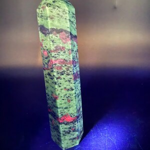 Op de afbeelding: Een hoge unakiet kristallen obelisk in groen en rood. De obelisk heeft een zeshoekige vorm en is gepolijst. De steen heeft een gespikkeld patroon van groen, rood en zwart. Bovenop zit een klein, lichtblauw kristal.