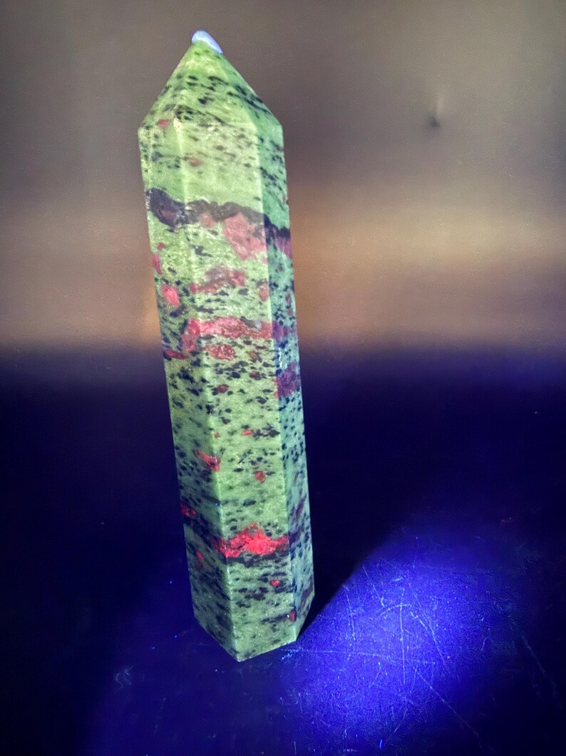 Op de afbeelding: Een hoge unakiet kristallen obelisk in groen en rood. De obelisk heeft een zeshoekige vorm en is gepolijst. De steen heeft een gespikkeld patroon van groen, rood en zwart. Bovenop zit een klein, lichtblauw kristal.