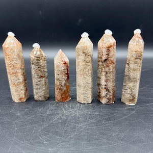 May include: Six natural crystal points with a mix of tan, brown, and reddish-brown hues. Each crystal has a unique pattern and is topped with a small, white, rounded cap. The crystals are arranged on a dark surface.
