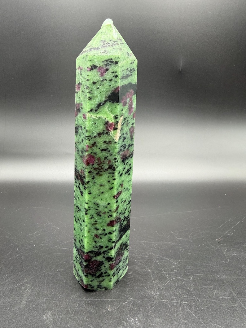 Op de afbeelding: Een hoge, gepolijste robijn-zoisiet kristallen toren. De steen is een levendig groen met zwarte en rode insluitsels. Het kristal heeft een facetvormige, zeshoekige vorm en een puntige top. De achtergrond is donkergrijs.