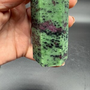 Op de afbeelding: Een groene en rode zoisiet kristallen toren. De gepolijste steen heeft een zeshoekige vorm en wordt in een hand gehouden. Het kristal heeft een gespikkeld patroon van zwarte en rode insluitsels door de groene basis.